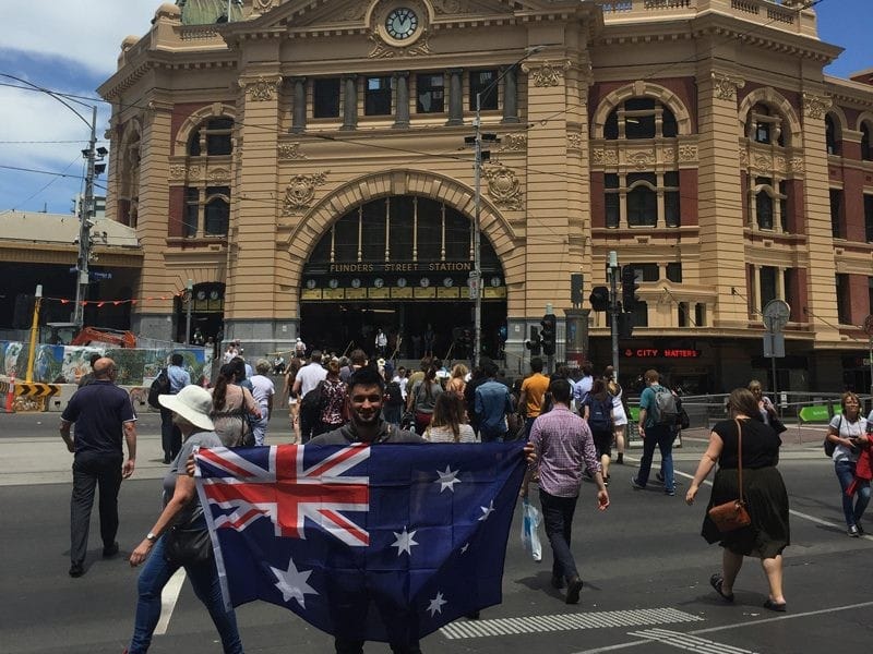 Vivir en Melbourne chico con la bandera de Australia en medio de la gente y enfrente de un monumento en la ciudad de Melbourne